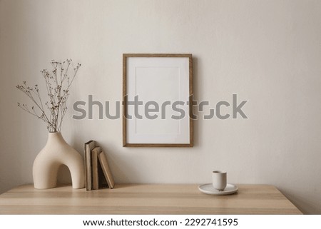 Empty wooden picture frame mockup hanging on beige wall background. Boho shaped vase, dry flowers on table. Cup of coffee, old books. Working space, home office. Art, poster display. Modern interior.