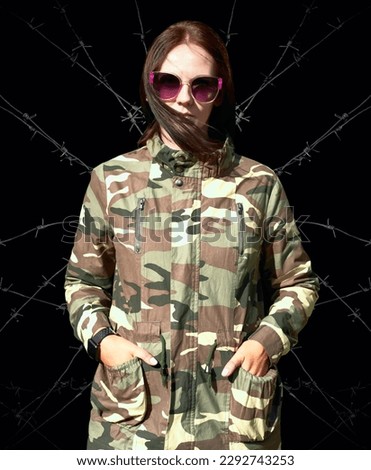 long haired girl wearing black sunglasses in olive khaki jacket looks at camera. stylish woman with big violet sunglasses. khaki pattern. isolated on black background. barbed wire behind female 
