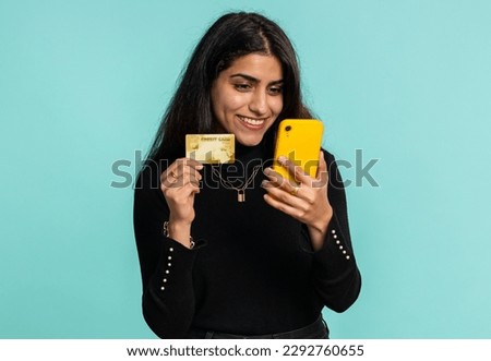 Sincere indian woman customer using credit bank card and smartphone while transferring money, purchases online shopping, payment. Finance and internet. Pretty girl isolated alone on blue background