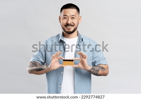 Attractive smiling Asian man wearing stylish casual clothes, holding credit card looking at camera isolated on gray background. Shopping, electronic money concept 