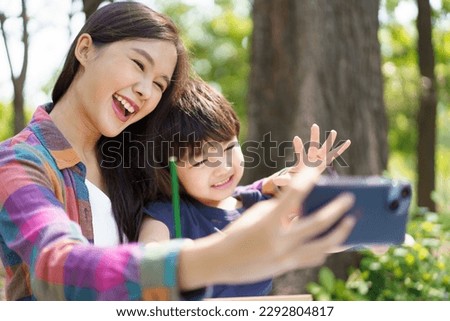 Happy cheerful Asian lovely little boy and mother sitting under the tree at park in the morning and drawing a picture and taking a selfie photography on smartphone together.