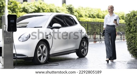 Businesswoman using tablet, walking while recharging her electric vehicle with charging station at public car parking. Progressive lifestyle of technology and ecological concern by EV car Royalty-Free Stock Photo #2292805189