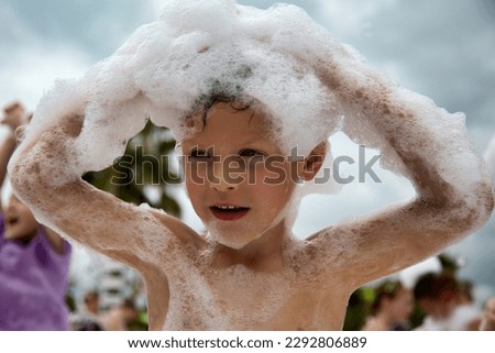 Cute funny boy with wet hands holds a lot of white foam on his head at a foam festival. Portrait of a Caucasian little boy having fun on the beach in white foam
