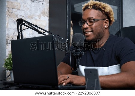 Smiling black guy in a black T-shirt and glasses, sitting in front of a laptop screen. A handsome man communicates online using a microphone and a laptop in a home studio