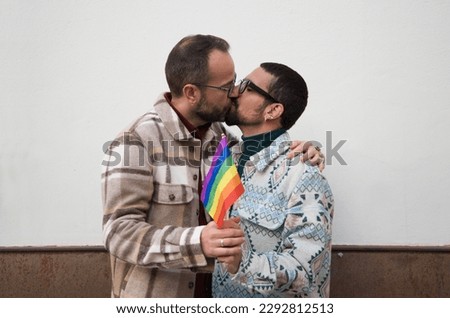 A young couple of gay men. The marriage is happy and they hug and kiss each other with the gay pride flag in their hands. Homosexual rights concept. Royalty-Free Stock Photo #2292812513