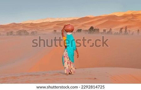 Woman in traditional green dress carrying heavy jug of water on her head and walking on the desert - Sahara, Morocco