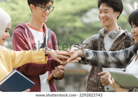 A group of diverse, multiracial young people are shown happily stacking their hands together while standing outside a building. This represents a sense of unity, fun,relationship and youth culture