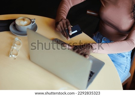 Cropped photo of African-American woman wearing pink sweater, sitting at table with cup of coffee in cafe, working, doing online shopping on laptop, holding credit card. E-commerce, internet banking.