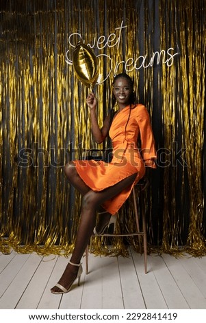 Attractive african american woman in elegant formal dress sit on chair in festive hall with gold decorations. Joyful gorgeous birthday woman in photo zone. Birthday party, showy holiday, celebration.
