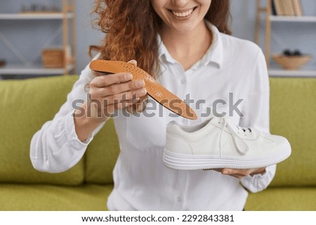 Cropped shot of happy, smiling young woman sitting on the sofa at home, holding a white orthopedic shoe and putting a new orthotic insole inside it. Footwear, comfort, feet health concept Royalty-Free Stock Photo #2292843381