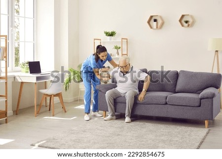 Young nurse in scrubs holding a weak retired old man by the hand and helping him stand up from the sofa at home. Senior care, treatment, rehabilitation, life in assisted living facility concept Royalty-Free Stock Photo #2292854675