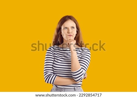Portrait of serious confused undecided young woman full of doubt holding hand on chin and thinking, trying to make hard choice decision. Girl with wavy hair in striped sweatshirt on yellow background. Royalty-Free Stock Photo #2292854717