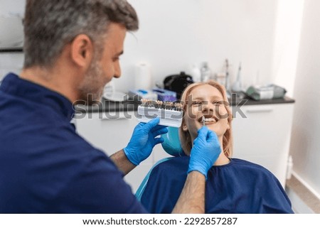 Image of satisfied young woman sitting in dental chair at medical center while professional doctor fixing her teeth, Female dentist choosing filling shade for smiling woman, using tooth scale sample