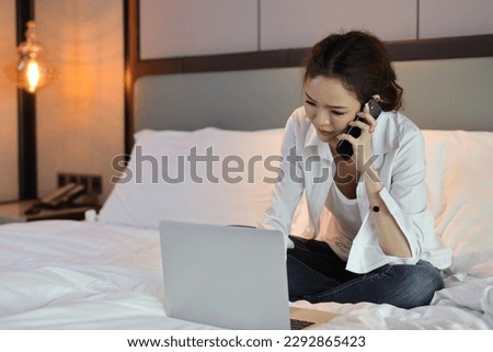 Active and unhappy asian woman in casual sitting on bed and working smart mobile phone and computer in hotel, beauty businesswoman with bedroom background. Lifestyle technology concept