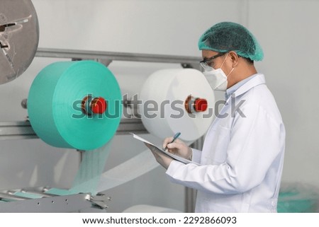 Worker man in personal protective equipment or PPE inspecting quality of mask and medical face mask production line in factory, manufacturing industry and factory concept.