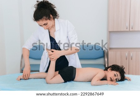 Confident female orthopedic doctor makes examination of little boy laying on massage table at medical office. Orthopedics, medical care, children health.