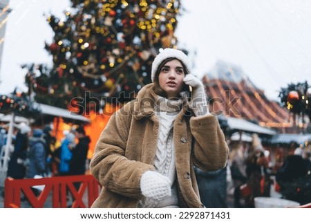 Trendy woman in warm coat and hat standing on street near decorated Christmas tree and having call on mobile phone