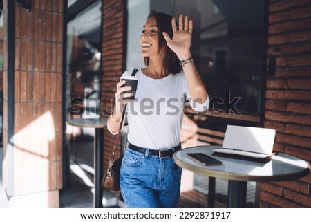 Elegant smiling female in casual clothes with cup of coffee standing at small table outside cafe looking away and waving hand Royalty-Free Stock Photo #2292871917