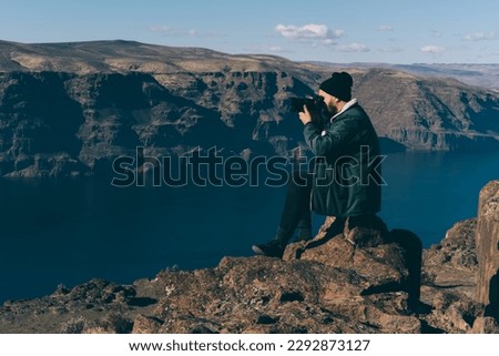 Side view of bearded male traveler sitting on rock near edge of cliff and taking pictures of amazing landscape of mountains and river