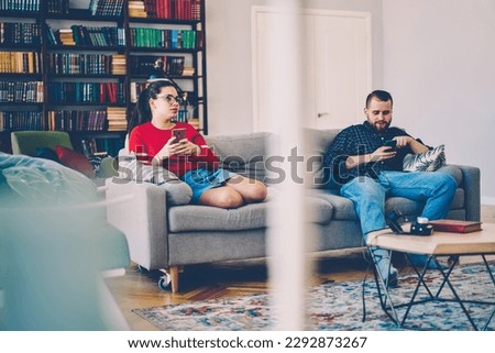 Tranquil couple in casual wear sitting on comfortable couch and browsing smartphones while chilling together during weekend in modern flat Royalty-Free Stock Photo #2292873267