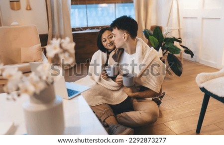 Multiracial couple sitting on floor with covering blanket at table at home at winter. Caucasian man and asian woman hold cups with tea or coffee and enjoy time together. Relationship and closeness Royalty-Free Stock Photo #2292873277