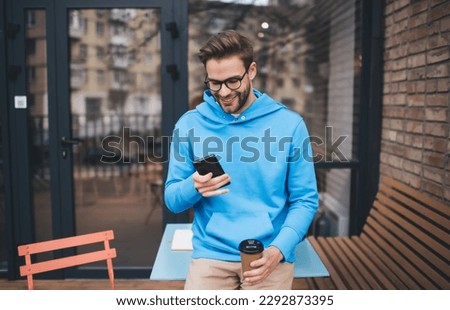 Millennial male in spectacles browsing website on modern cellphone device using 4g wireless during coffee break at street terrace, happy Caucaisan hipster guy in optical eyewear chatting and texting Royalty-Free Stock Photo #2292873395