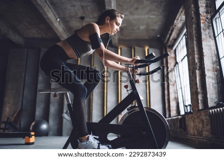 Side view of young fit female with device on arm and earphones doing cardio practice on stationary exercise bicycle in gym Royalty-Free Stock Photo #2292873439