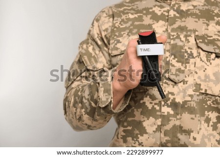 Soldier in military uniform holding medical tourniquet on light grey background, closeup. Space for text