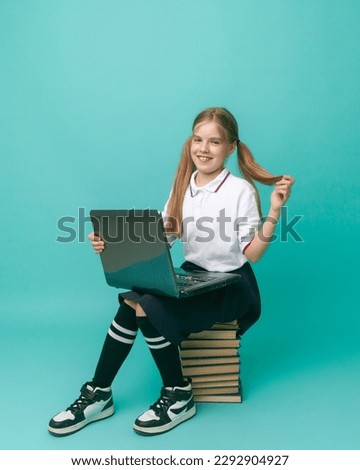 a schoolgirl is sitting on a stack of books with a laptop, on a blue background in the studio. Learning concept
