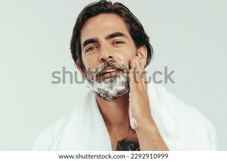 Young man appliying shaving cream to his beard, preparing himself for a smooth and comfortable shave. Man practicing his grooming routine, taking care of his facial hair to maintain a smooth look. Royalty-Free Stock Photo #2292910999