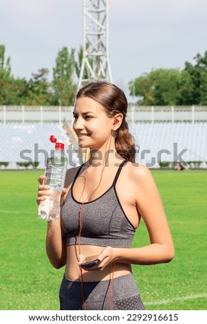 Young girl drinking water from bottle after running at stadium. Sports and healthy concept. woman holding phone with music.