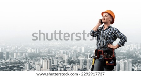 Attractive blonde woman in workwear and hardhat talking on smartphone. Portrait of young worker in checkered blue shirt standing on cityscape background. Business communication and conversation.