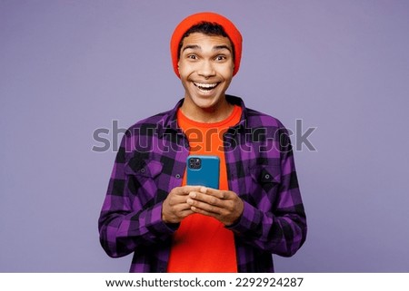 Young overjoyed excited happy man of African American ethnicity wear casual shirt orange hat hold in hand use mobile cell phone isolated on plain pastel light purple color background studio portrait