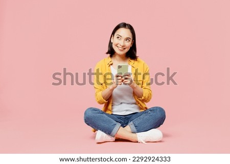 Full body young woman of Asian ethnicity wear yellow shirt white t-shirt sitting hold in hand use mobile cell phone isolated on plain pastel light pink background studio portrait. Lifestyle concept Royalty-Free Stock Photo #2292924333