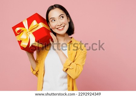 Young minded woman of Asian ethnicity wear yellow shirt white t-shirt hold in hand present box with gift ribbon bow look aside isolated on plain pastel light pink background studio. Lifestyle concept. Royalty-Free Stock Photo #2292924349