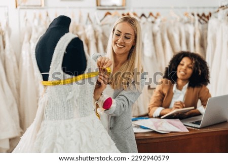 A beautiful woman is measuring chest circumference of a bridal dress while her colleague writes measures Royalty-Free Stock Photo #2292940579