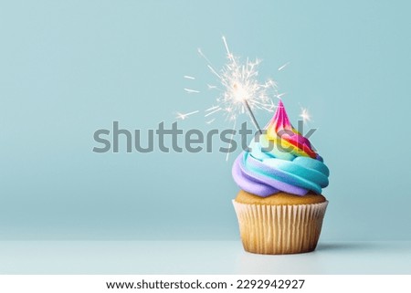 Birthday cupcake with colorful rainbow frosting and celebration sparkler for a birthday party, plain blue background with copy space to side
