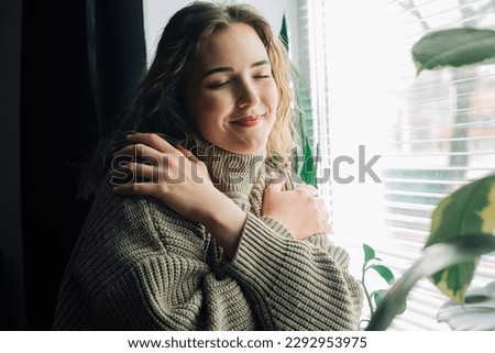 Thoughtful woman standing next to window, looking through, hugging herself, feeling lonely waiting for her boyfriend or dreaming of true love and healthy relationship. Royalty-Free Stock Photo #2292953975