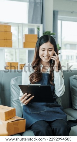Make mobile phone call, Young pretty Asian female influencer small businesses SME owners female entrepreneurs working on receipt box and check online orders, sitting on sofa of home online business.