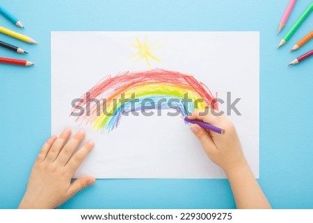 Little child hand holding pencil and drawing colorful rainbow on white paper on light blue table background. Pastel color. Closeup. Point of view shot. Children creativity time.