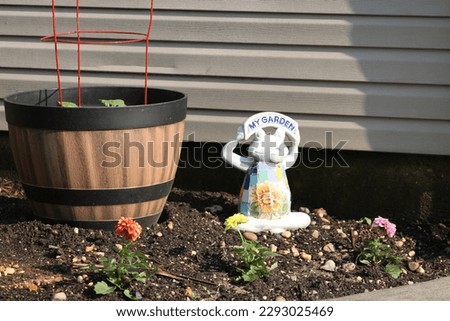 A frog statue with a sign that says "My Garden" on it. It's sitting in the dirt near potted vegetable plants and dahlia flowers.