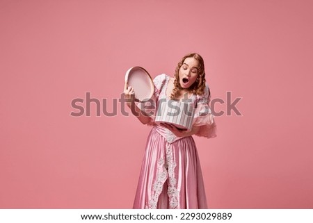 Pleasant surprise. Portrait with happy, beautiful, blond princess wearing pink dress and holding gift box with surprised face on pink studio background. Medieval, beauty, birthday, emotions concept