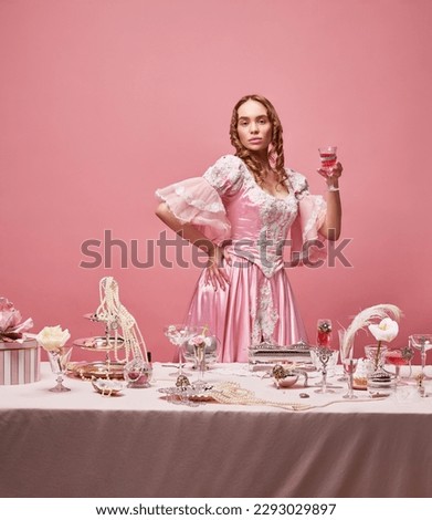Serious, arrogant queen. Shot of beautiful woman, princess wearing renaissance pink dress and standing with wine glass over studio background. Concept of medieval, human emotions, beauty, fashion, ad