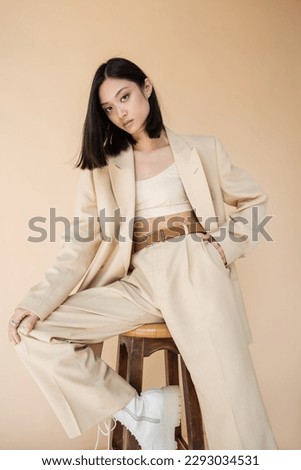 stylish asian woman in pantsuit holding hand in pocket while posing on stool isolated on beige Royalty-Free Stock Photo #2293034531