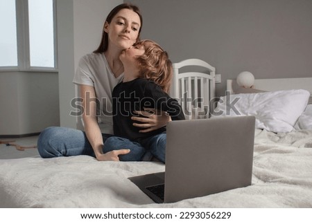 Happy mother with her little son enjoying in online shopping or working from home. Business from distance and virtual communication. Mom and baby watch cartoons or play game on laptop on the bed.