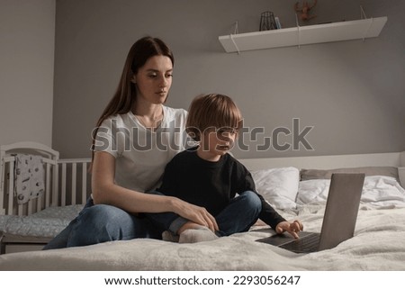 Happy mother with her little son enjoying in online shopping or working from home. Business from distance and virtual communication. Mom and baby watch cartoons or play game on laptop on the bed.