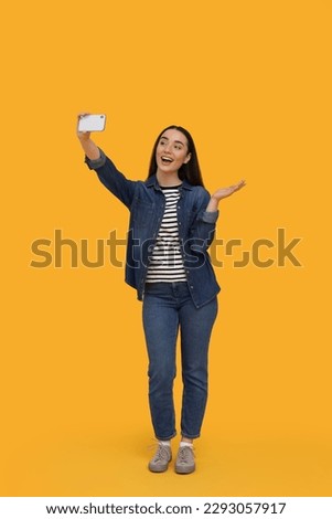 Smiling young woman taking selfie with smartphone on yellow background