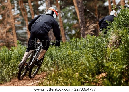 rear view male racer riding on forest trail downhill race, black sports clothes, biking high plants Royalty-Free Stock Photo #2293064797