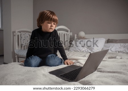 Little boy sitting on bed and looking at laptop. The kid watches cartoons and plays on the computer. 
Toddler learns something new and interesting.