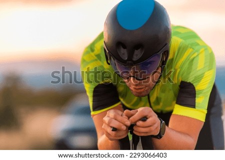  Close up photo of triathlete riding his bicycle during sunset, preparing for a marathon. The warm colors of the sky provide a beautiful backdrop for his determined and focused effort. Royalty-Free Stock Photo #2293066403